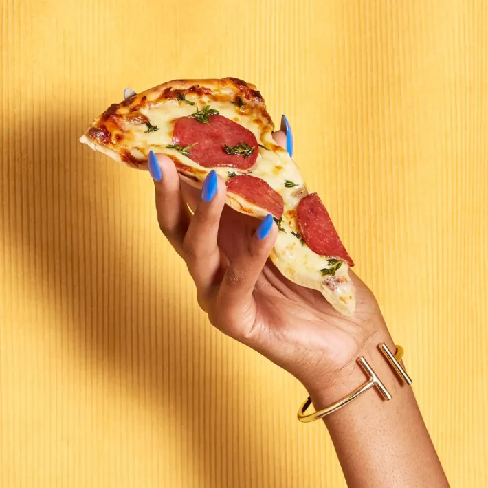 Greasy Glamour Content styled hands with bright finger nails holding slice off pizza in front of velvet background